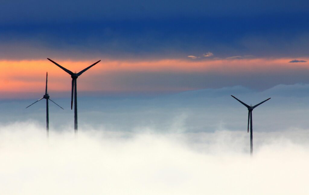 Silhouetted wind turbines rise above clouds against a dramatic sunset sky.